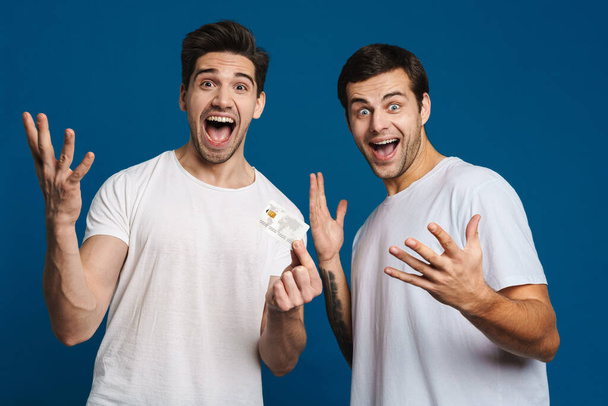 Excited two guys expressing surprise while posing with credit card isolated over blue background - Фото, изображение