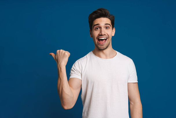 Excited unshaven guy smiling and pointing finger aside isolated over blue background - Fotó, kép