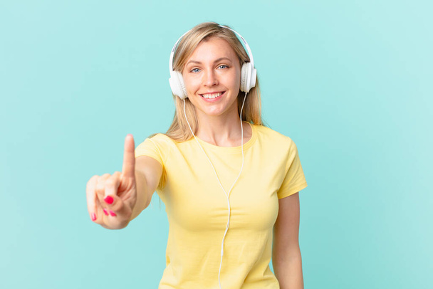 young blonde woman smiling and looking friendly, showing number one and listening music. - Foto, Imagem