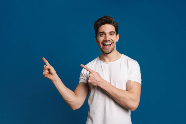 Happy unshaven guy smiling and pointing fingers aside isolated over blue background - Фото, изображение
