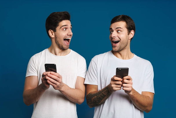 Очарованные два парня выразили удивление, позируя со смартфонами, изолированными на синем фоне - Фото, изображение