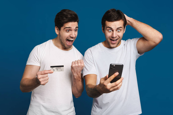 Delighted two guys posing with credit card and mobile phone isolated over blue background - Foto, imagen