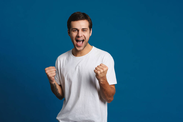 Excited unshaven guy screaming and making winner gesture isolated over blue background - 写真・画像