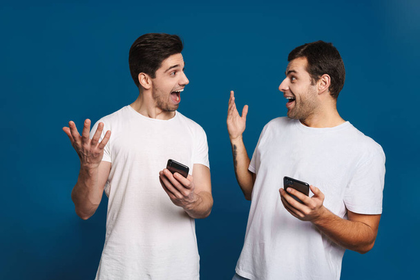 Delighted two guys expressing surprise while posing with smartphones isolated over blue background - Foto, Bild