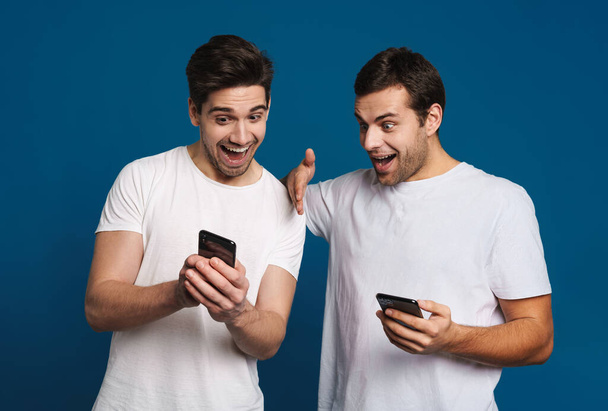 Delighted two guys expressing surprise while posing with smartphones isolated over blue background - Foto, Bild
