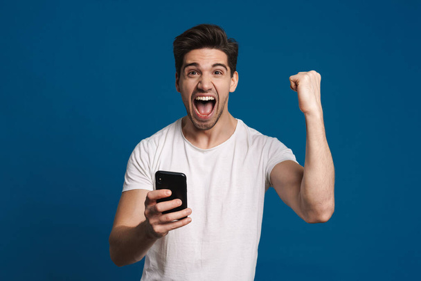 Delighted guy making winner gesture while using mobile phone isolated over blue background - Foto, Imagen