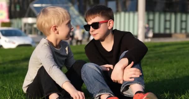 Children lively discuss something. Two boys on the lawn in sunglasses have fun chatting. Brothers on rest on the lawn - Footage, Video