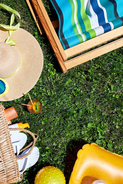 Flat lay of outdoor bright summer beach vacation or travel lifestyle concept with straw bag, hat, flip flops, lemonade, inflatable yellow mattress and ball, sunglasses and sunscreen spray on grass background, top view, copy space. - Photo, Image