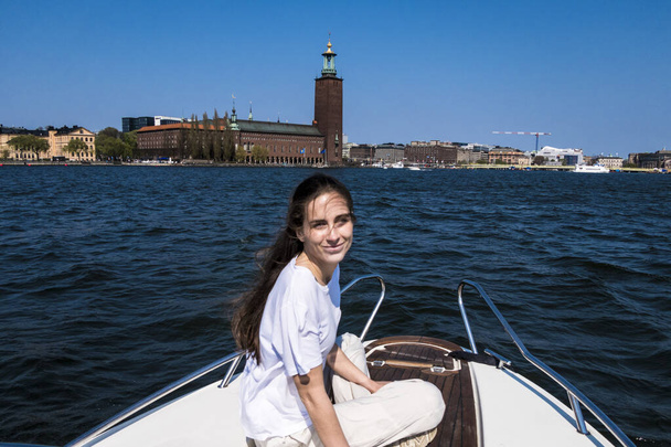 Stockholm, İsveç 'te genç bir kadın arka planda CIty Hall olan bir motorlu teknenin pruvasında oturuyor.. - Fotoğraf, Görsel