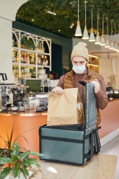 Vertical portrait of delivery man wearing mask while packing takeout orders to bag in cafe interior - Foto, Imagem