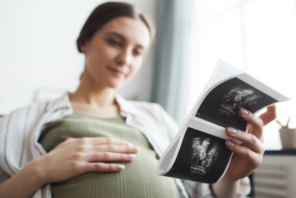 Mujer embarazada sosteniendo la imagen de rayos X de su bebé durante su descanso en el sofá en casa - Foto, imagen