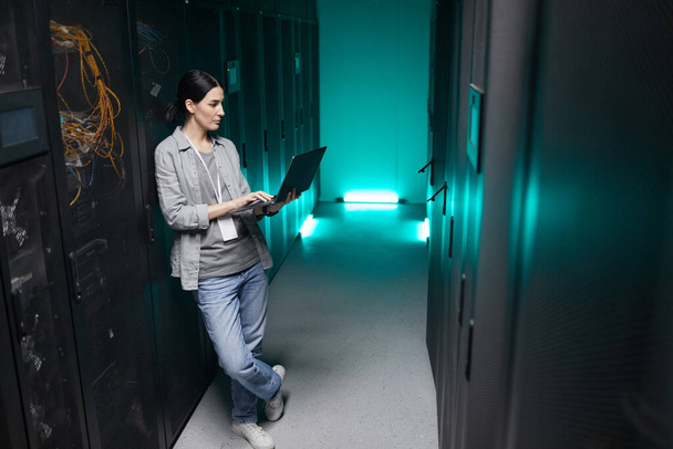 Retrato de ángulo alto de longitud completa del ingeniero de datos femenino que sostiene el ordenador portátil mientras trabaja con la supercomputadora en la sala de servidores, espacio de copia - Foto, imagen