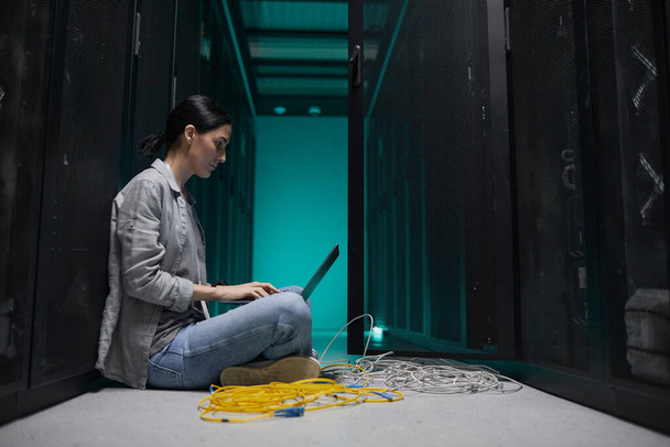 Retrato de vista lateral de una mujer ingeniera de datos usando una computadora portátil mientras está sentada en el piso en la sala de servidores y configurando una red de supercomputadoras, espacio para copiar - Foto, imagen