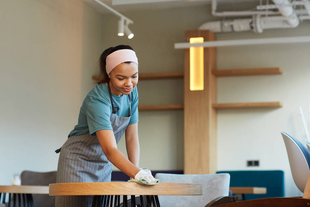 Portrait of smiling mixed-race waitress cleaning table in cafe or coffee shop while preparing for opening in morning, copy space - Photo, Image
