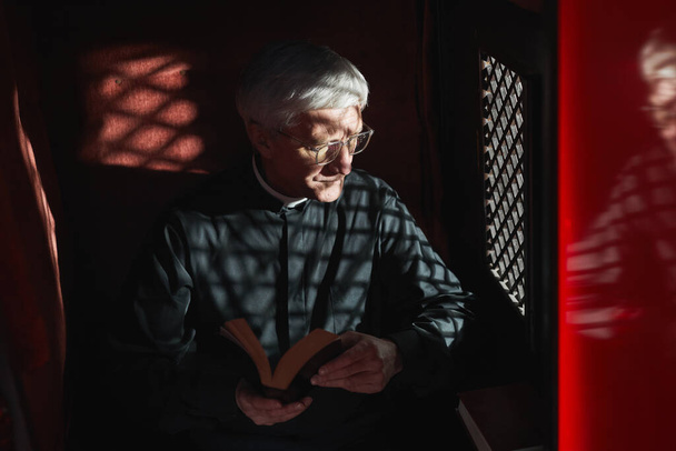 Sacerdote anziano seduto con la Bibbia in confessionale e in ascolto della confessione - Foto, immagini