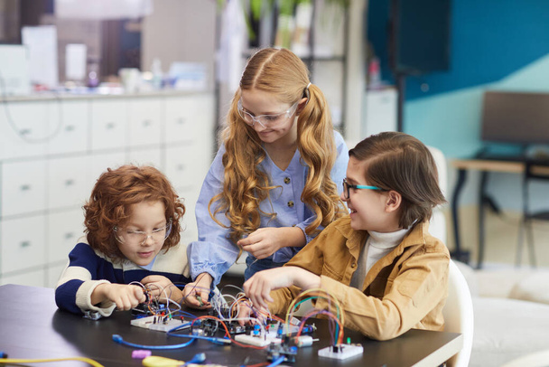 Portret trójki uśmiechniętych dzieci eksperymentujących z obwodami elektrycznymi podczas budowy robotów w klasie inżynierskiej w szkole - Zdjęcie, obraz