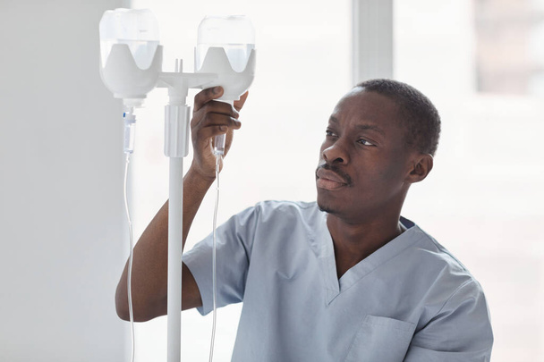 Afro-Amerikalı erkek hemşirenin belden portresi. Hastanedeki hastaları tedavi ederken serum serumu takıyor. - Fotoğraf, Görsel