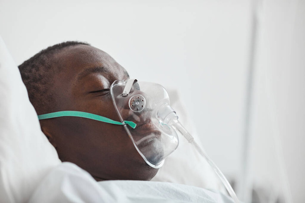 Zijaanzicht portret van jonge Afro-Amerikaanse man in ziekenhuisbed met zuurstofmasker, kopieerruimte - Foto, afbeelding