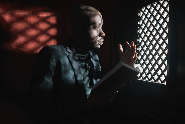 Afrikaanse jonge priester die de Bijbel leest terwijl hij in de biechtstoel zit - Foto, afbeelding