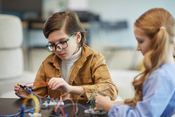 Retrato de un chico lindo usando gafas y experimentando con circuitos eléctricos mientras construye robots durante la clase de ingeniería en la escuela - Foto, imagen