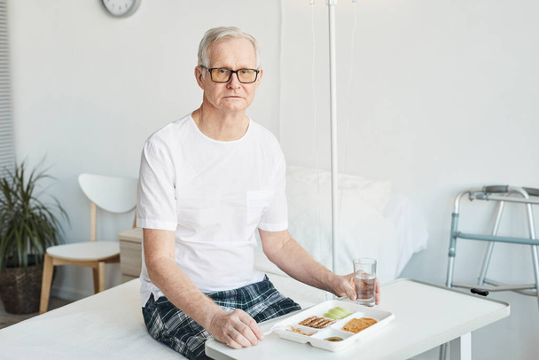 Portret van een oudere man die eten eet in de ziekenhuiskamer en naar camera kijkt, ruimte kopieert - Foto, afbeelding