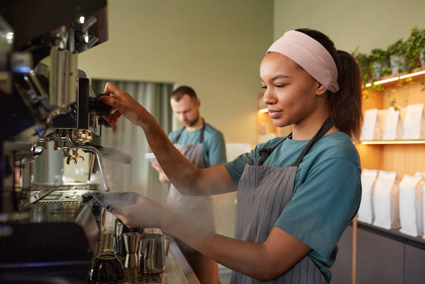 Side view portrait of young female barista making fresh coffee in cafe and operating coffee machine - Foto, afbeelding