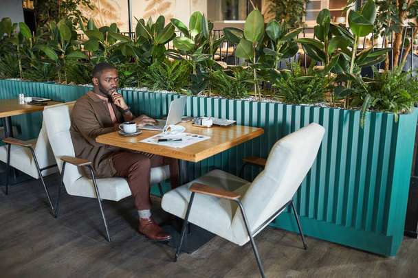 Teljes hosszúságú portré sikeres afro-amerikai üzletember dolgozik laptop, miközben ül az asztalnál környezetbarát zöld kávézó belsejében, másolja a helyet - Fotó, kép