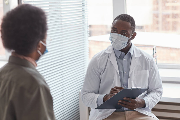Afrykański lekarz w masce ochronnej rozmawia ze swoim pacjentem podczas wizyty w szpitalu - Zdjęcie, obraz