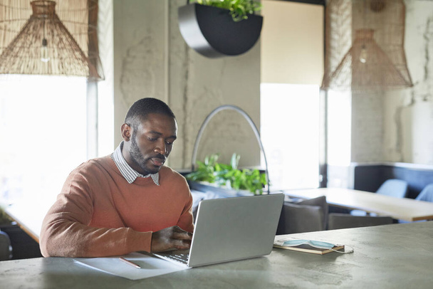 Portrait of adult African-American man using laptop while working in modern cafe interior decorated with fresh green plants, copy space - 写真・画像