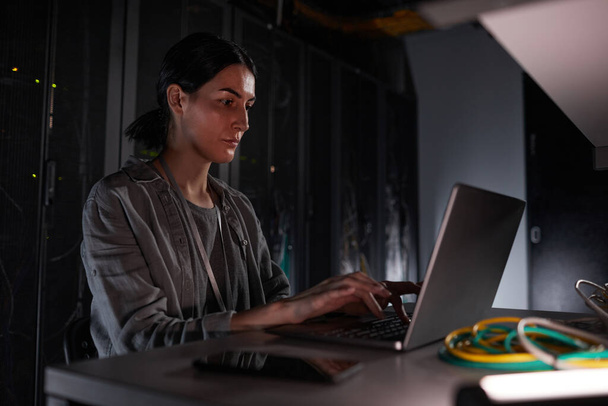 Retrato de la mujer ingeniero de red utilizando el ordenador portátil mientras está sentado en la habitación del servidor oscuro, espacio de copia - Foto, Imagen