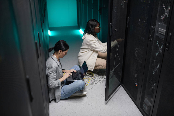 サーバールームのラップトップを使用した2人の女性データエンジニアの肖像とスーパーコンピュータネットワークのセットアップ、コピースペース - 写真・画像