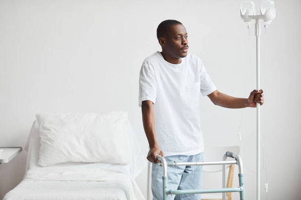 Portret van een Afro-Amerikaanse man in het ziekenhuis die een infuusstandaard vasthoudt en gepensioneerd wegkijkt, kopieert ruimte. - Foto, afbeelding