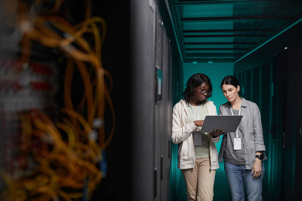 Retrato de gran angular de dos mujeres jóvenes que utilizan el ordenador portátil en la sala de servidores mientras se configura la red de supercomputadoras, espacio de copia - Foto, imagen