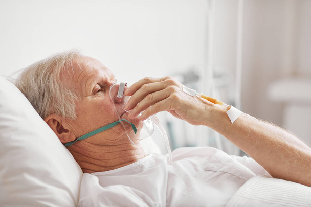 Вид сбоку портрет больного пожилого человека лежащего в больничной койке с кислородной маской и капельницей, пространство для копирования - Фото, изображение