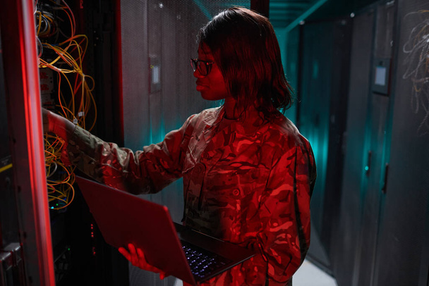 Oldalnézet portré afro-amerikai nő irányító számítógépes hálózat, miközben dolgozik a szuperszámítógép szerver szobában világít a piros fény, másolás tér - Fotó, kép