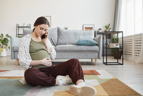 Schwangere sitzt auf dem Boden und telefoniert im Wohnzimmer - Foto, Bild