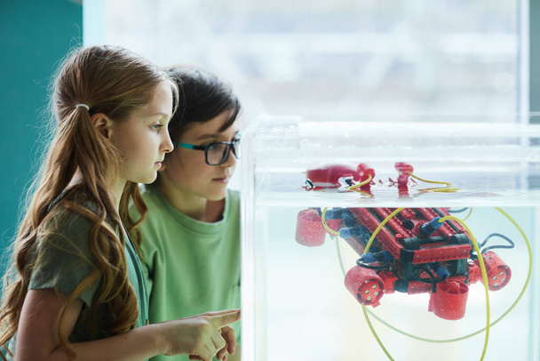Retrato de vista lateral de dos niños operando un bote robótico en un tanque de agua mientras experimentan en un laboratorio de ingeniería en la escuela, espacio para copiar - Foto, Imagen