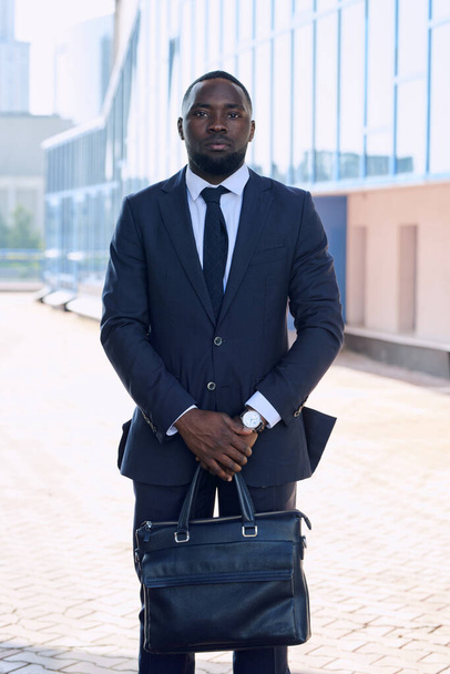 Молодой уверенный в себе африканский бизнесмен в формальной одежде стоит перед камерой напротив внешнего вида современного здания в городской среде - Фото, изображение