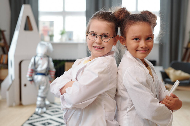 Portrait of two little girls in medical coats standing back to back at home and smiling at camera - Photo, Image
