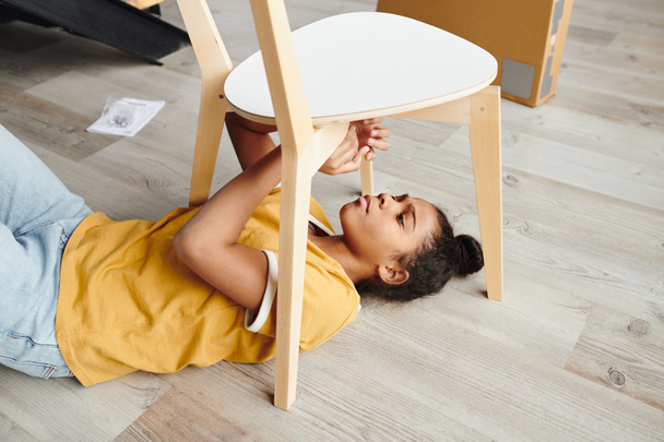 Милая девочка-подросток смешанной расы в повседневной одежде собирает деревянный стул, лежа на полу гостиной и чинит сиденье - Фото, изображение