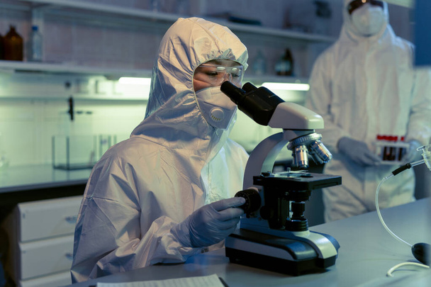 Cientista em vestuário de trabalho de proteção examinando amostras através do microscópio na mesa no laboratório químico - Foto, Imagem