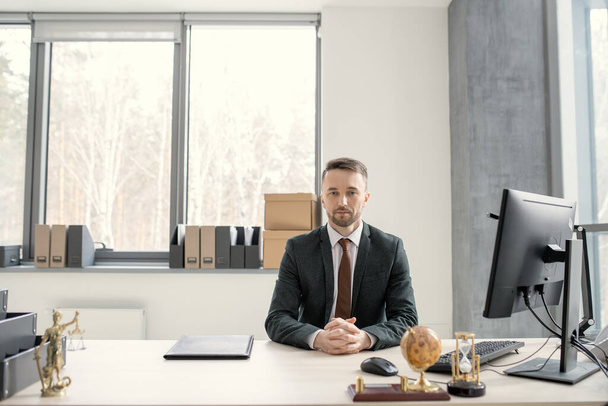 Retrato de joven ejecutivo guapo serio en traje negro y corbata sentado con las manos apretadas en la mesa equipada con computadora - Foto, imagen