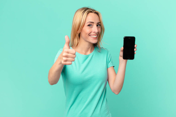 young blonde woman feeling proud,smiling positively with thumbs up and holding a smart phone - Фото, изображение