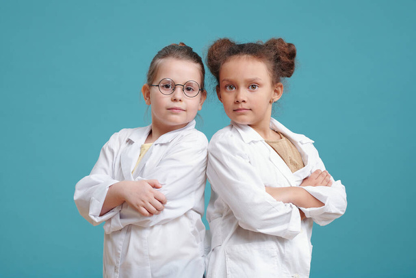 Portrait of two little girls in white coats standing with arms crossed and looking at camera against the blue background - Fotoğraf, Görsel