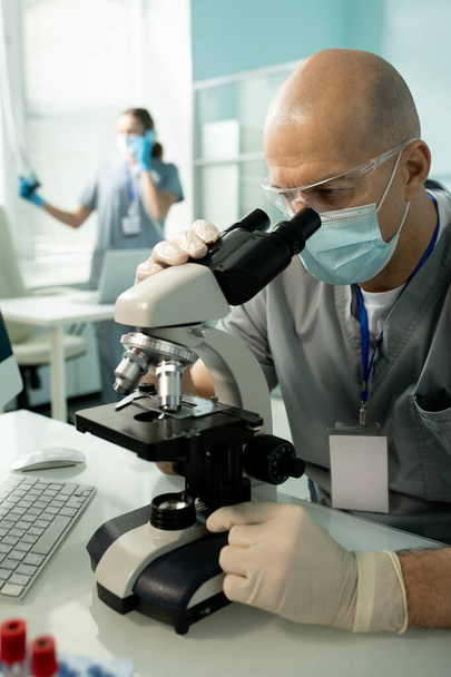 Empleado de laboratorio calvo concentrado en gafas y guantes de látex sentado en la mesa y trabajando con el microscopio mientras investigaba - Foto, imagen