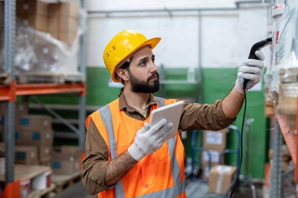 Серьезный молодой бородатый мужчина в каске и перчатках сканирует код товара, делая инвентаризацию запасов на складе - Фото, изображение