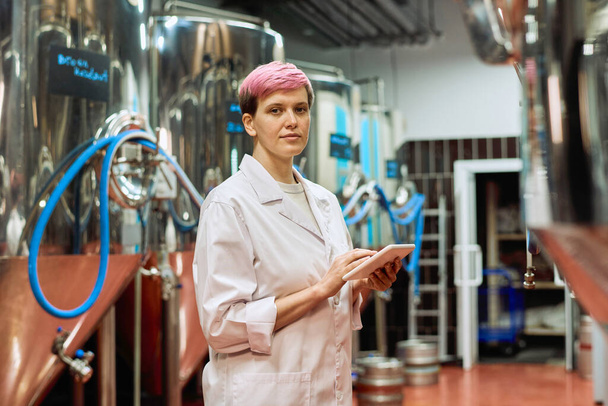 Jonge serieuze vrouwelijke brouwerij expert in whitecoat kijken naar u tijdens het gebruik van digitale tablet tegen nieuwe apparatuur voor bierproductie in de fabriek - Foto, afbeelding