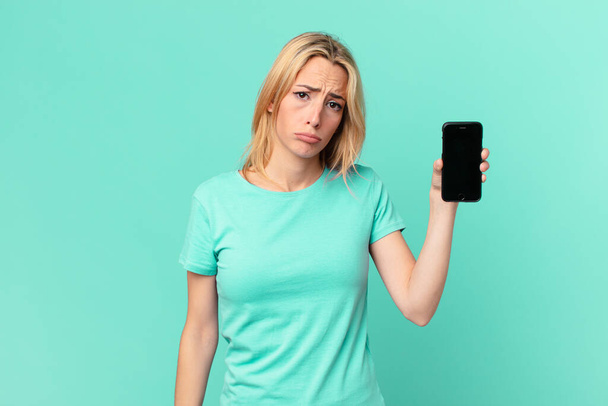 young blonde woman feeling sad and whiney with an unhappy look and crying and holding a smart phone - Fotó, kép