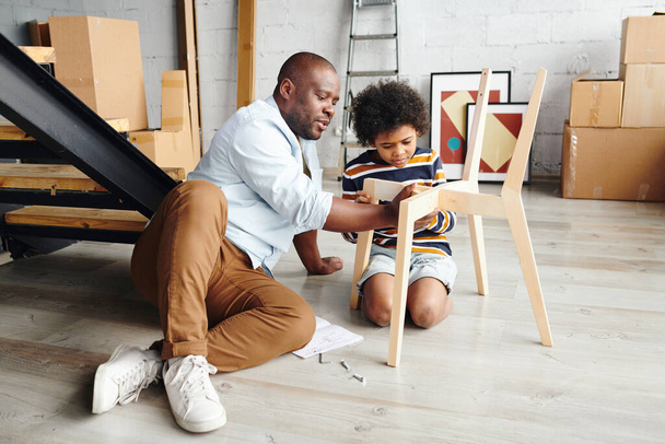 Giovane padre in casualwear insegnare al suo piccolo figlio carino come assemblare sedia in legno durante il fissaggio delle sue parti, entrambi seduti sul pavimento - Foto, immagini