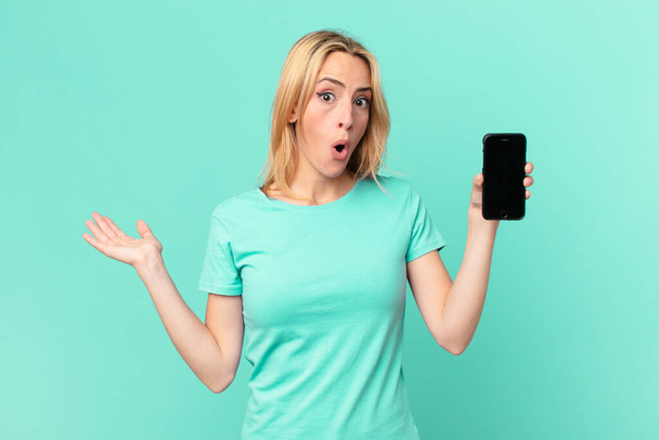 young blonde woman looking surprised and shocked, with jaw dropped holding an object and holding a smart phone - Photo, Image